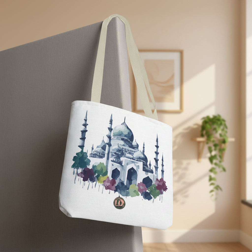 A white canvas tote bag with beige shoulder straps, featuring a watercolor-style illustration of a mosque with minarets and colorful flowers, shown hanging against a neutral gray background.