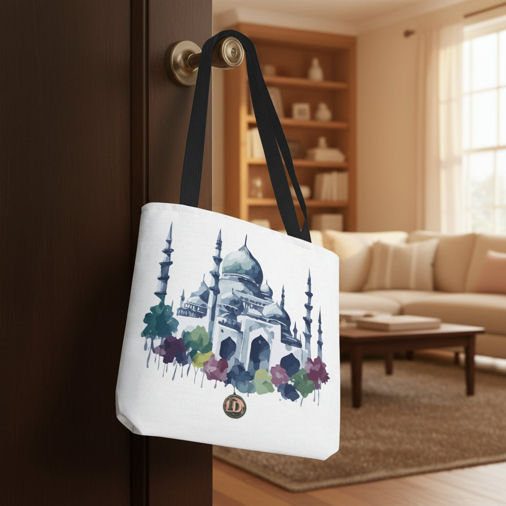 A white canvas tote bag with black shoulder straps, featuring a watercolor-style illustration of a mosque with blue, green, and purple floral accents, hanging against a neutral gray background.