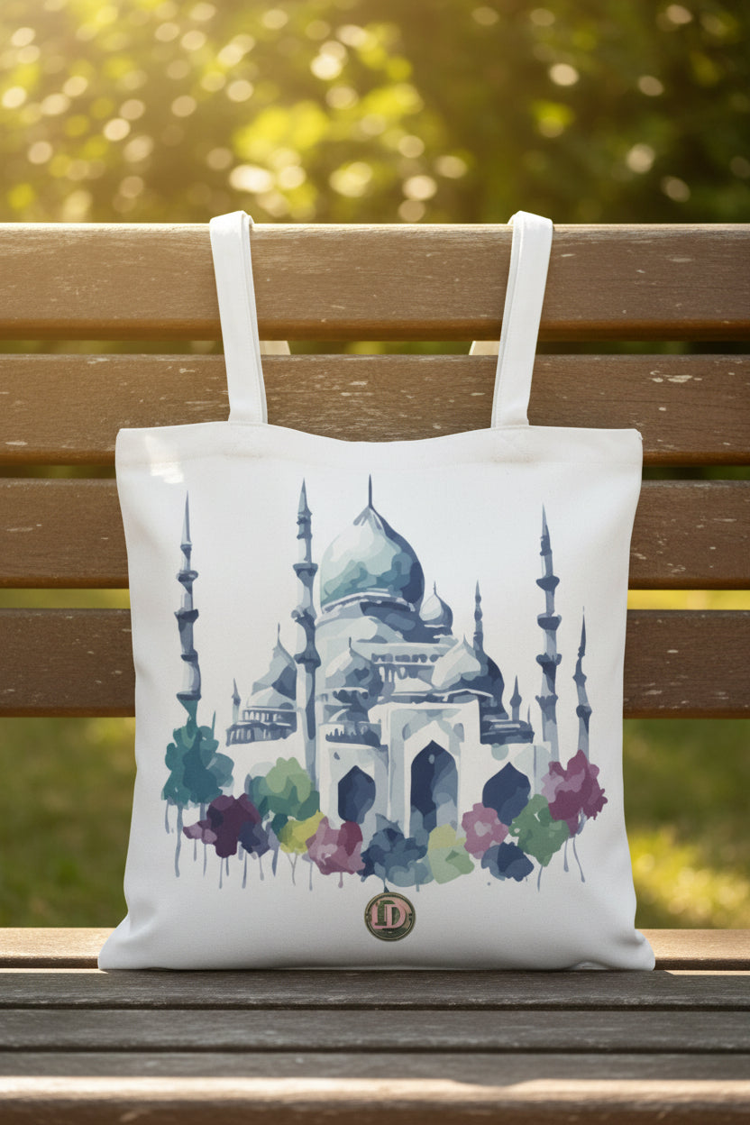 A white canvas tote bag with a watercolor-style illustration of a mosque and colorful trees, featuring two shoulder straps and a small circular logo near the bottom.