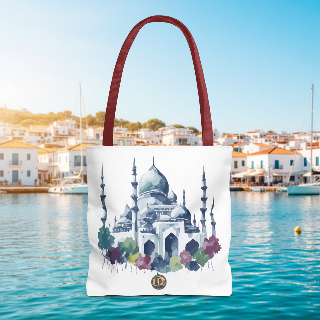 A white canvas tote bag with dark red shoulder straps, featuring a watercolor-style illustration of a mosque with blue, green, and purple tones, and a small circular monogram logo near the bottom.