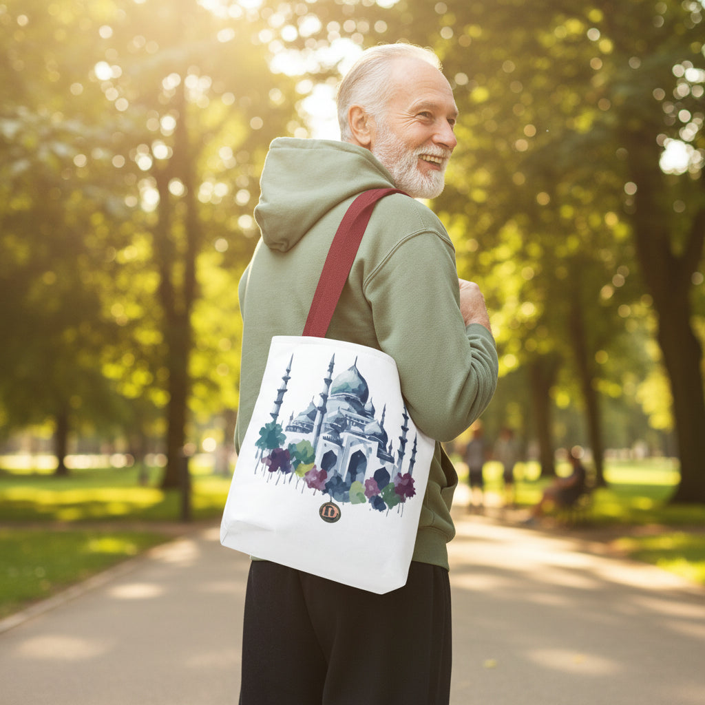 A man wearing an olive green hoodie and dark pants is shown from the back and side, carrying a white canvas tote bag over his shoulder. The bag has a red strap and a printed illustration of a mosque with blue, purple, and green watercolor details.