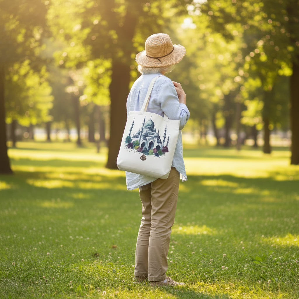 An older lady wearing an olive green hoodie and dark pants is shown from the back and side, carrying a white canvas tote bag over her shoulder. The tote bag features a watercolor-style illustration of a domed mosque surrounded by trees and colorful floral accents, with a small circular gold emblem near the bottom. She is in the sunny park