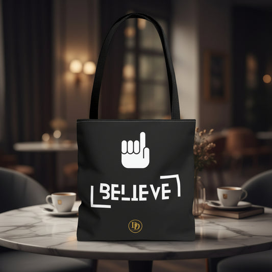 A black canvas tote bag with black shoulder straps, featuring a white hand graphic and the word 'BELIEVE' in white, with a small gold circular 'LD' logo near the bottom. Muslim tote bag.