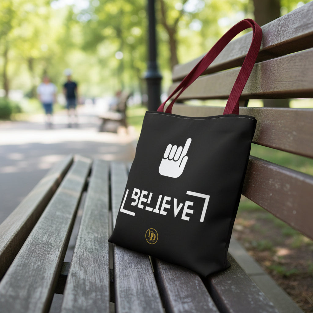 A black tote bag with dark red shoulder straps, featuring a white hand graphic and the word 'BELIEVE' printed on the front, with a small gold circular 'DD' logo below. Muslimah tote bag. 